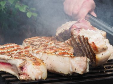 Siyah önlüklü bir aşçı kemiklerinde biftek pişirir. Kemikte ızgara domuz eti. Izgara için maşa.