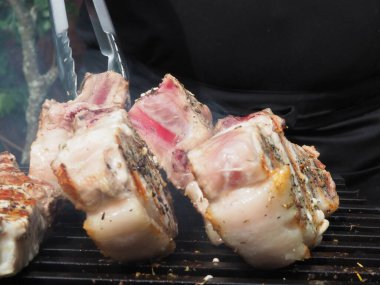 Siyah önlüklü bir aşçı kemiklerinde biftek pişirir. Kemikte ızgara domuz eti. Izgara için maşa.