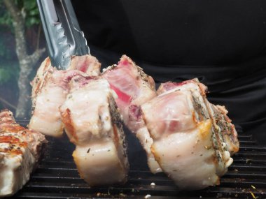 Siyah önlüklü bir aşçı kemiklerinde biftek pişirir. Kemikte ızgara domuz eti. Izgara için maşa.