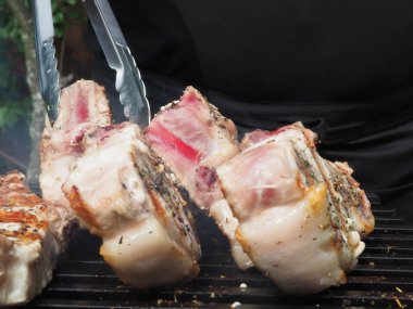 Siyah önlüklü bir aşçı kemiklerinde biftek pişirir. Kemikte ızgara domuz eti. Izgara için maşa.