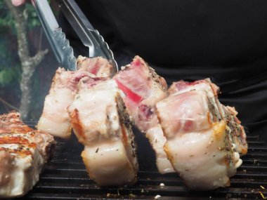 Siyah önlüklü bir aşçı kemiklerinde biftek pişirir. Kemikte ızgara domuz eti. Izgara için maşa.