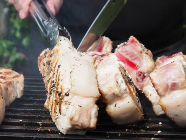 Siyah önlüklü bir aşçı kemiklerinde biftek pişirir. Kemikte ızgara domuz eti. Izgara için maşa.