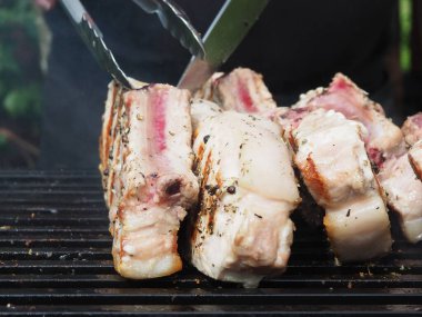 Siyah önlüklü bir aşçı kemiklerinde biftek pişirir. Kemikte ızgara domuz eti. Izgara için maşa.