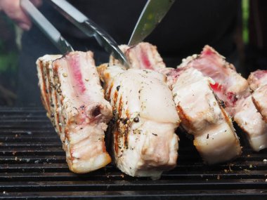 Siyah önlüklü bir aşçı kemiklerinde biftek pişirir. Kemikte ızgara domuz eti. Izgara için maşa.