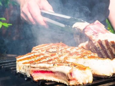 Adam biftekleri maşayla çeviriyor. Kömürde pişirmek. Kemiklerdeki domuz eti.