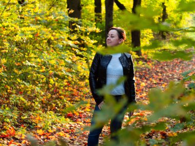 Altın sonbahar parkında deri ceketli genç bir kadın. Parka düşen yapraklar.