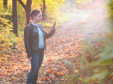 Parkta deri ceketli genç bir kadın selfie çekiyor. Parkta sonbahar