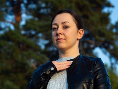 Deri ceketli genç bir kadın ceketini düzeltiyor. Yeşil bir arka planda. Temiz havada yürüyorum..