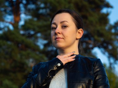 Deri ceketli genç bir kadın ceketini düzeltiyor. Yeşil bir arka planda. Temiz havada yürüyorum..