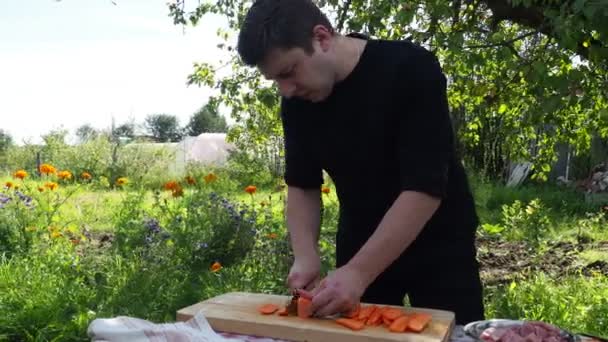 El hombre está cortando zanahorias en una junta en el área suburbana. un joven con una chaqueta negra prepara verduras — Vídeos de Stock