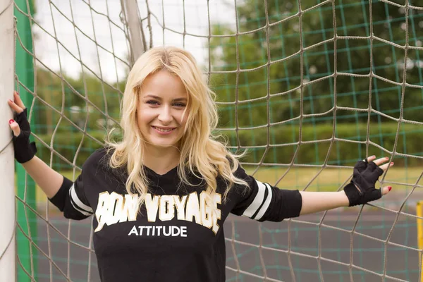 A beautiful blonde at the football net is holding and smiling during the game.