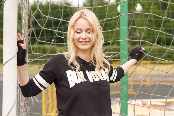 A beautiful blonde at the football net is holding and smiling during the game.