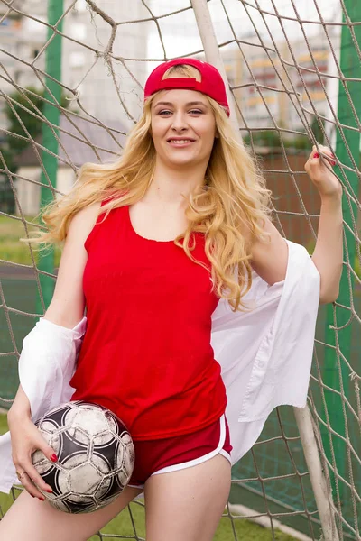 The blonde in red form with a ball at the gate on the football field. Photo shoot at the football goal.