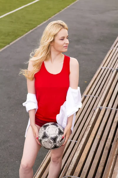 Girl football player stands with the ball at the bench in red. Posing for a photographer on the edge of a football field.