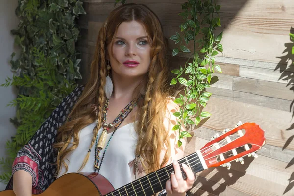 stock image A beautiful girl with red curly hair stands and holds a seven-string guitar in her hands. Studio with green leaves.