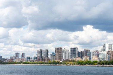The scene captures a vibrant city skyline beside a calm waterfront, with tall buildings reaching for the sky under a blanket of clouds. Green trees add a touch of nature to the urban environment.