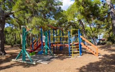 Parlak renkli oyun alanı ekipmanları uzun ağaçlarla çevrili gölgeli bir alanda duruyor. Aileler kumsalın yakınındaki güneşli günün tadını çıkarıyorlar. Çocuklar için mükemmel bir ortam yaratıyorlar..