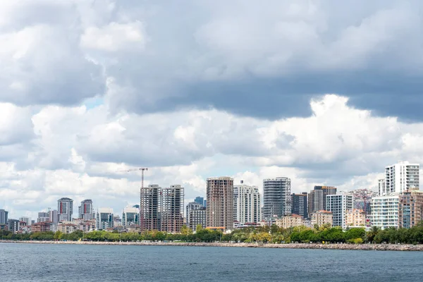 The scene captures a vibrant city skyline beside a calm waterfront, with tall buildings reaching for the sky under a blanket of clouds. Green trees add a touch of nature to the urban environment.