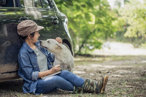 Meyve bahçesinde arabasının yanında oturan ve köpeğinin kafasını okşayan Asyalı genç bir kadın..