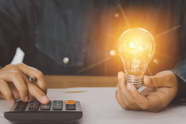 İnsan eli bir ampulü tutuyor ve şirketin enerji kullanım rakamlarını hesaplamak için hesap makinesine basıyor. Enerji ve muhasebe kavramının korunması.