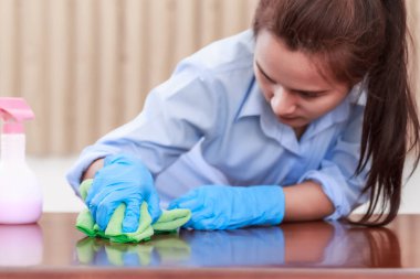 Mavi koruyucu eldivenler giyen bir kadın elinde yeşil bir mikrofiber bez tutarak ahşap masaları temizliyor ve iyi hijyen için dezenfekte, temizlik ve dezenfekte yöntemleri içeren sprey şişeleri sıkıyor..