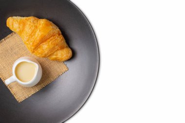 Siyah tabakta kruvasan ve beyaz arka planda izole edilmiş yoğunlaştırılmış süt ve çuval bezi. Kırpma yolu ile. Fransız kruvasanı kahvaltısı. Üst görünüm.
