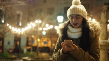 Beautiful smiling girl messaging with smartphone on the winter street with christmas lights in SLOW MOTION HD VIDEO. Glowing blurred Christmas lights decoration and falling snow in the background.
