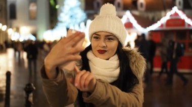 Beautiful smiling girl making selfie with smartphone on the winter street with christmas lights in SLOW MOTION HD VIDEO. Glowing blurred Christmas lights decoration in the background.