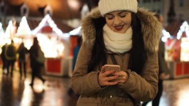 Beautiful smiling girl messaging with smartphone on the winter street with christmas lights in SLOW MOTION HD VIDEO. Glowing blurred Christmas lights decoration in the background. Close-up. Half Speed.