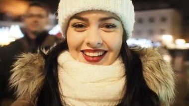 Beautiful smiling girl making selfie centrifuge with smartphone on the winter street with christmas lights in SLOW MOTION HD VIDEO. Glowing blurred Christmas lights decoration in the background.