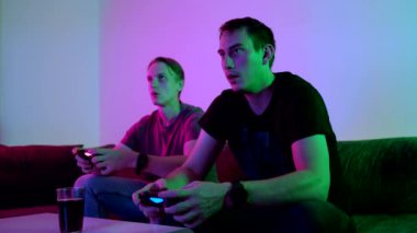 Two happy friends playing action video game in the living room sitting on the sofa in 4K VIDEO. Intense competition between two young male players is full of emotions. 