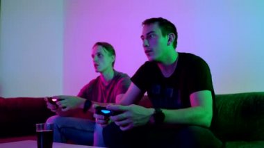 Two happy friends playing action video game in the living room sitting on the sofa in 4K VIDEO. Intense competition between two young male players is full of emotions. 