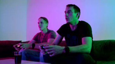 Two happy friends playing action video game in the living room sitting on the sofa in 4K VIDEO. Intense competition between two young male players is full of emotions. 
