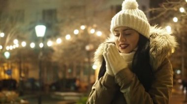 Young happy woman is warming up her gloved hands in cold winter evening on street in SLOW MOTION HD VIDEO. She is smiling. Snowing, glowing blurred Christmas lights and walking people in background. Half speed.