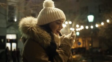 Young happy woman is warming up her gloved hands in cold winter evening on street in SLOW MOTION HD VIDEO. She is smiling. Snowing, glowing blurred Christmas lights and walking people in background. Half speed.