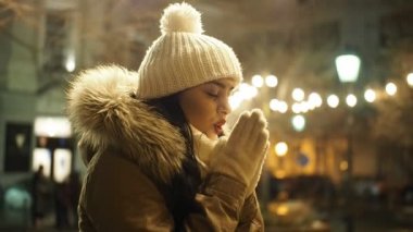 Young happy woman is warming up her gloved hands in cold winter evening on street in SLOW MOTION HD VIDEO. She is smiling. Snowing, glowing blurred Christmas lights and walking people in background. Half speed.