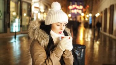 Young happy woman is drinking hot punch in cold winter evening on street in SLOW MOTION HD VIDEO. She is smiling. Glowing blurred Christmas lights and walking people in background. Half speed.