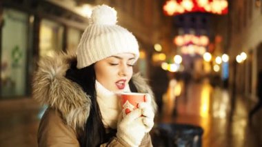 Young happy woman is drinking hot punch in cold winter evening on street in SLOW MOTION HD VIDEO. She is smiling. Glowing blurred Christmas lights and walking people in background. Half speed.
