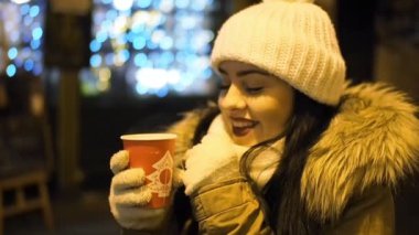 Young happy woman is drinking hot punch in cold winter evening on street in SLOW MOTION HD VIDEO. She is smiling. Glowing blurred Christmas lights and walking people in background. Half speed.