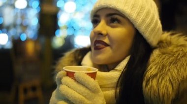 Young happy woman is drinking hot punch in cold winter evening on street in SLOW MOTION HD VIDEO. She is smiling. Glowing blurred Christmas lights and walking people in background. Half speed.