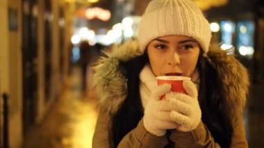 Young happy woman is drinking hot punch in cold winter evening on street in SLOW MOTION HD VIDEO. She is smiling. Glowing blurred Christmas lights and walking people in background. Half speed.