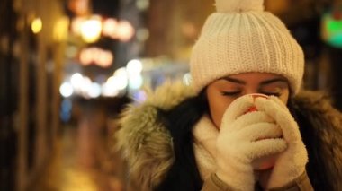 Young happy woman is drinking hot punch in cold winter evening on street in SLOW MOTION HD VIDEO. She is smiling. Glowing blurred Christmas lights and walking people in background. Half speed.