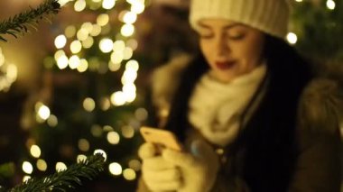 SLOW Motion HD VIDEO 'da ışıklandırılmış Noel ağacı ile cep telefonuyla mesajlaşan güzel bir kız. Arka planda parlak, bulanık ışıklar var. Yakın plan. Yarı Hız.