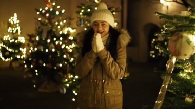 SLOW Motion HD VIDEO 'da aydınlatılmış Noel ağacı tarafından ısıtılan güzel kız. Arka planda parlak, bulanık ışıklar var. Yakın plan. Yarı Hız.