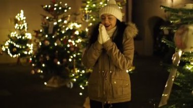 SLOW Motion HD VIDEO 'da aydınlatılmış Noel ağacı tarafından ısıtılan güzel kız. Arka planda parlak, bulanık ışıklar var. Yakın plan. Yarı Hız.