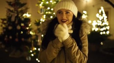 SLOW Motion HD VIDEO 'da aydınlatılmış Noel ağacı tarafından ısıtılan güzel kız. Arka planda parlak, bulanık ışıklar var. Yakın plan. Yarı Hız.