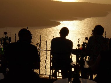 Terastaki geleneksel bir kafede dinlenen insanların siluetleri batan güneşin güzel turuncu ışınlarının arka planına karşı. Günbatımında Caldera ve deniz.