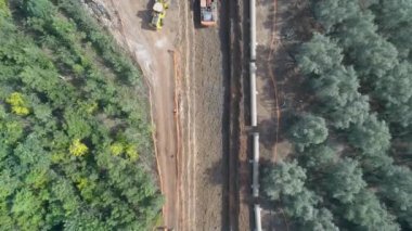 İnşaat alanının dışındaki yeşil ağaçlarla birlikte hava aracı. Müteahhit vinçle kazı yapıyor ve bir su kanalını kürekliyor beton boru hatları yerleştiriyor ve yerleştiriyor.