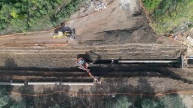 İnşaat alanının dışındaki yeşil ağaçlarla birlikte hava aracı. Müteahhit vinçle kazı yapıyor ve bir su kanalını kürekliyor beton boru hatları yerleştiriyor ve yerleştiriyor.