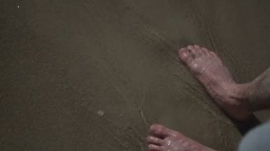 Yürüyen, yürüyen ve deniz sahilindeki altın kumda duran bir adam. Süper yavaş çekim. Okyanus kıyısında yürürken erkek bacakları alçak açıyla çekiliyor. Yalınayak dalgalarla kumlu sahilde. Yaz kıyı şeridi.